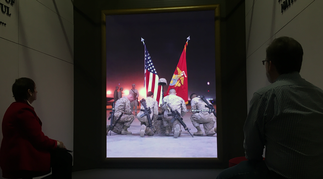 Two visitors watch large screen video with marines kneeling at a fallen marine memorial.